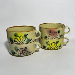 May include: Four stacked ceramic soup cups with handles. The cups are a pale yellow with a brown rim and feature a hand-painted floral design in yellow, pink, and green. The cups are stacked in pairs, with two sets visible.