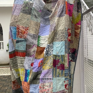 May include: A patchwork quilt with a variety of colors and patterns, including floral, geometric, and abstract designs. The quilt is made of recycled saris and is hand-stitched. The quilt is hanging on a wall and has the text "WOMENKNACKS" printed on it.