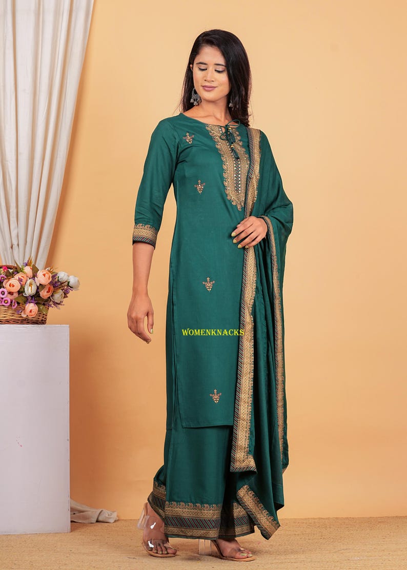 May include: A teal green kurta with gold embroidery and a matching palazzo pant. The kurta has a tie-up neckline and the palazzo pant has a gold border. The outfit is paired with a matching teal green dupatta with gold embroidery.