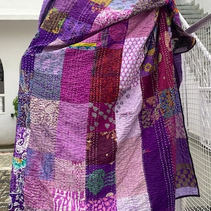 May include: A purple patchwork quilt with a variety of floral and geometric patterns. The quilt is made of recycled saris and is hand-stitched. The quilt is hanging on a tree branch and has the text "WOMEN KNACKS" printed on the bottom.