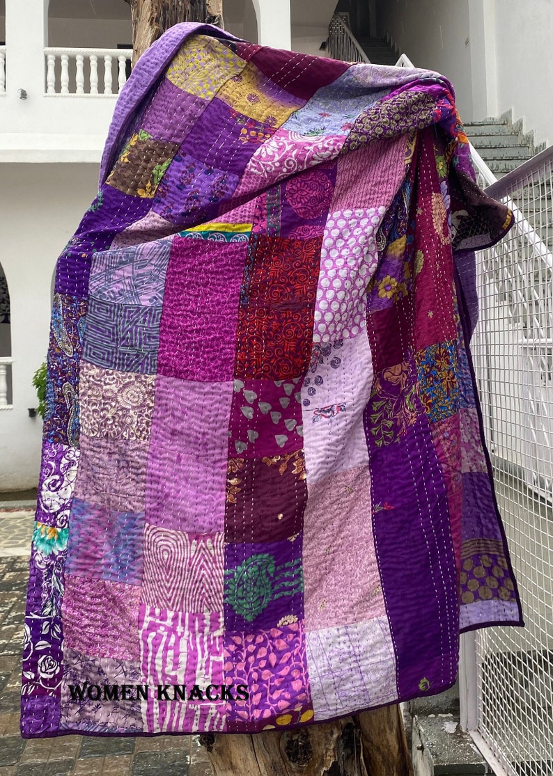 May include: A purple patchwork quilt with a variety of floral and geometric patterns. The quilt is made of recycled saris and is hand-stitched. The quilt is hanging on a tree branch and has the text "WOMEN KNACKS" printed on the bottom.