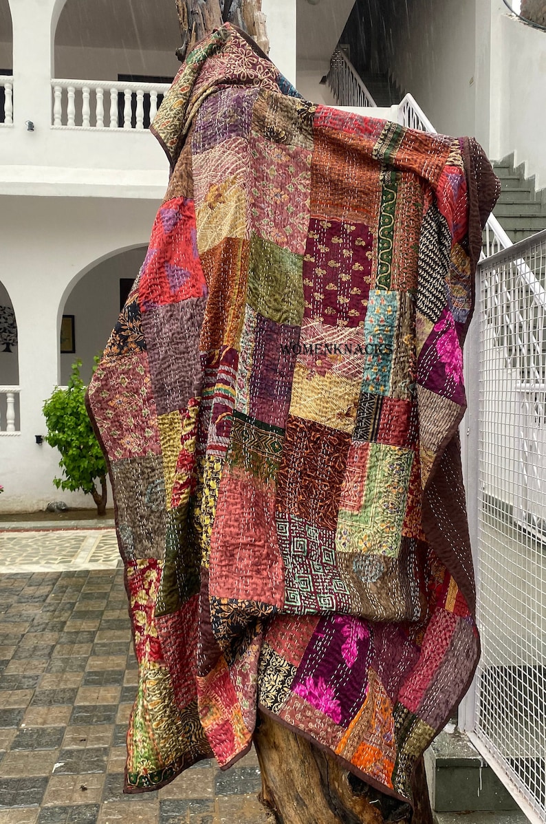 May include: A multi-colored patchwork quilt made from recycled saris. The quilt is a mix of brown, red, pink, green, and blue fabrics. The quilt is draped over a tree branch.