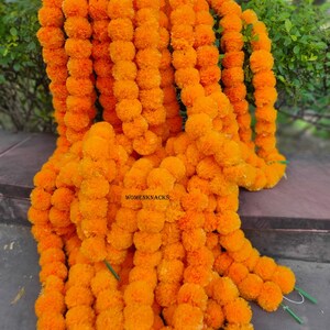 Può includere: Ghirlande di fiori di calendula arancioni brillanti. Le ghirlande sono fatte di molti piccoli fiori rotondi e soffici. I fiori sono infilati in lunghi fili. Il testo "WOMENKNACKS" è visibile sull'immagine.
