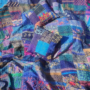 May include: A patchwork quilt and two matching throw pillows in shades of blue, purple, and green. The quilt and pillows are made of various fabric scraps, stitched together with visible white thread. The design features floral and geometric patterns.