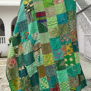 May include: A green patchwork quilt with a variety of patterns and colors. The quilt is made of recycled saris and is hand-stitched. The quilt is hanging on a tree trunk and has the text "Women Knacks" printed on the bottom.