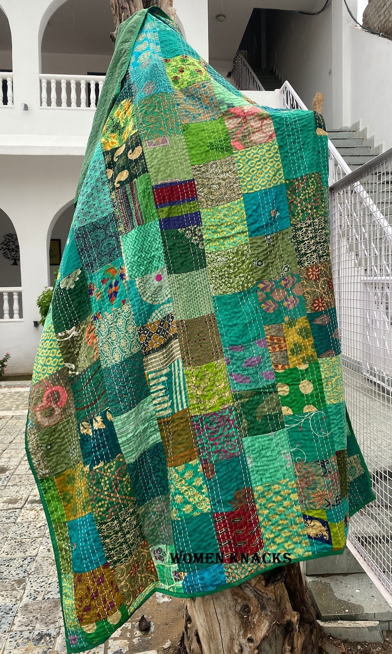 May include: A green patchwork quilt with a variety of patterns and colors. The quilt is made of recycled saris and is hand-stitched. The quilt is hanging on a tree trunk and has the text "Women Knacks" printed on the bottom.