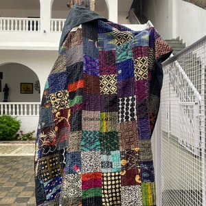 Peut inclure: Une courtepointe patchwork noire et multicolore avec une variété de motifs et de textures. La courtepointe est suspendue à une branche d'arbre devant un bâtiment blanc avec un escalier.