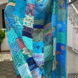 Pu&ograve; includere: Una trapunta patchwork caratterizzata da una variet&agrave; di quadrati di tessuto blu, verde acqua e turchese con diversi motivi floreali, paisley e geometrici. Il tessuto &egrave; cucito con ricami kantha bianchi e riporta il testo WOMENKNACKS in nero.