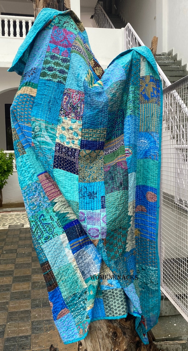 Pu&ograve; includere: Una trapunta patchwork caratterizzata da una variet&agrave; di quadrati di tessuto blu, verde acqua e turchese con diversi motivi floreali, paisley e geometrici. Il tessuto &egrave; cucito con ricami kantha bianchi e riporta il testo WOMENKNACKS in nero.