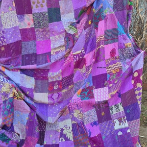 Könnte beinhalten: Eine große violette Patchwork-Decke aus verschiedenen Stoffen mit unterschiedlichen Mustern. Die Decke besteht aus Quadraten in verschiedenen Größen und Farben, und der Stoff ist mit einem Steppstich zusammengenäht. Die Decke ist in der Mitte gefaltet und über einen Ast gelegt.