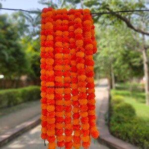 May include: A garland of bright orange marigold flowers hangs against a blurred outdoor background. The flowers are densely packed, creating a full, textured appearance. The garland is suspended from a thin wire, showcasing the warm colours of the blooms.