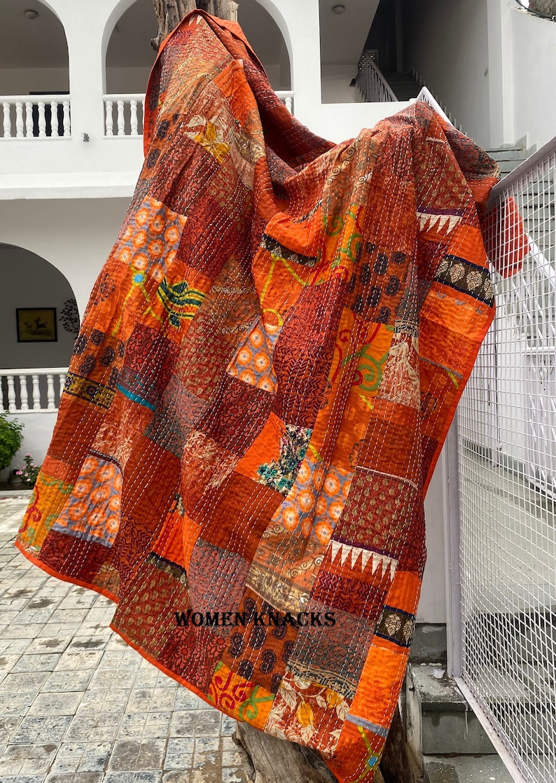 May include: A large, orange and brown patchwork quilt with intricate stitching. The quilt is made of various fabrics with floral and geometric patterns. The quilt is draped over a wooden post and has the text "WOMEN KNACKS" printed on the bottom right corner.