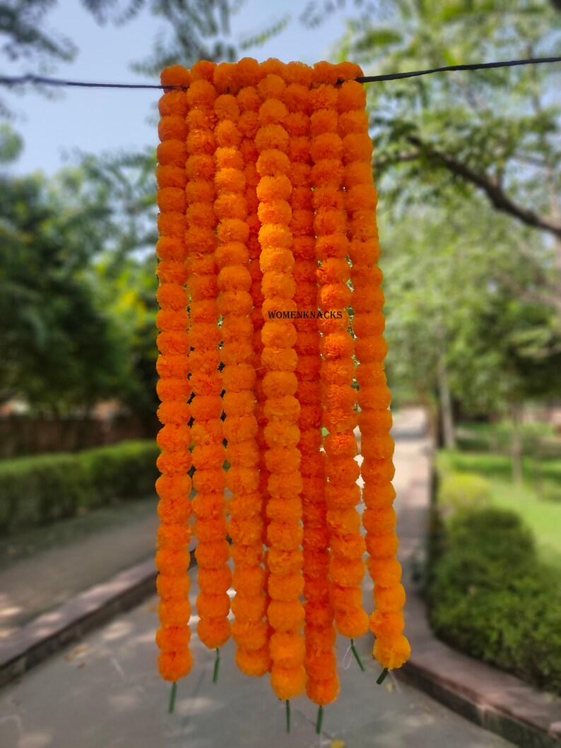 K&ouml;nnte beinhalten: Eine Girlande aus leuchtend orangefarbenen Ringelblumenbl&uuml;ten, die auf einem Draht aufgereiht sind. Die Girlande h&auml;ngt an einem Draht und ist scharf vor einem verschwommenen Hintergrund aus gr&uuml;nen B&auml;umen und einem gepflasterten Weg.