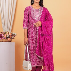 May include: A magenta kurta with a white floral print and a matching magenta dupatta with a white and yellow geometric print. The kurta has a round neckline and three-quarter sleeves. The dupatta is draped over the shoulder and has a gold border. The model is wearing a white handbag and clear heels.