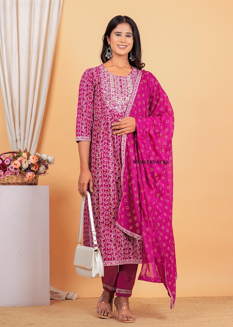 May include: A magenta kurta with a white floral print and a matching magenta dupatta with a white and yellow geometric print. The kurta has a round neckline and three-quarter sleeves. The dupatta is draped over the shoulder and has a gold border. The model is wearing a white handbag and clear heels.