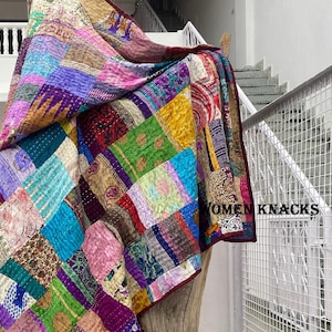 May include: A colorful patchwork quilt made from repurposed fabric scraps. The quilt is a mix of different colors, patterns, and textures, including purple, pink, blue, green, and brown. The quilt is draped over a wooden surface and has the text "YOMEN KNACKS" printed on the bottom right corner.