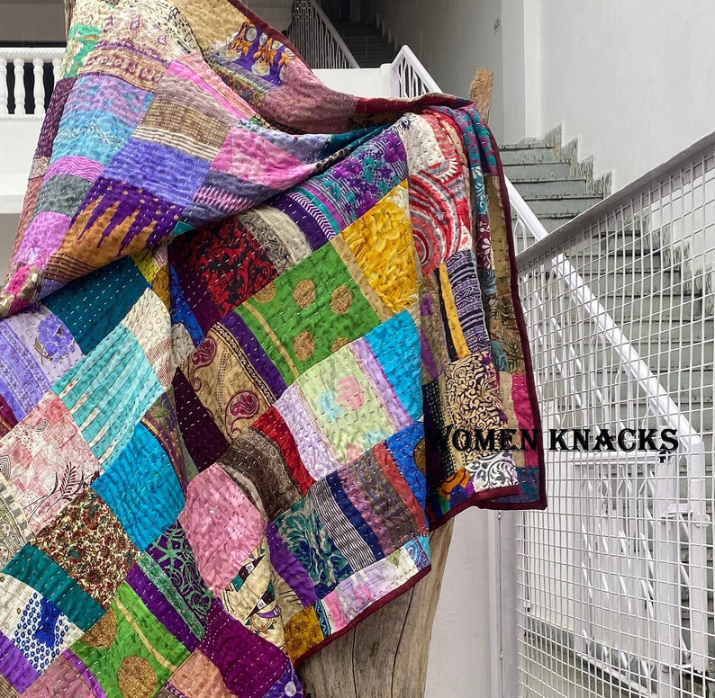 May include: A colorful patchwork quilt made from repurposed fabric scraps. The quilt is a mix of different colors, patterns, and textures, including purple, pink, blue, green, and brown. The quilt is draped over a wooden surface and has the text "YOMEN KNACKS" printed on the bottom right corner.