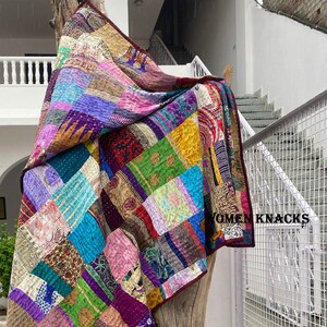 Pu&ograve; includere: Una trapunta patchwork composta da vari quadrati di tessuto colorato con motivi diversi, inclusi disegni floreali, paisley e geometrici. La trapunta presenta cuciture kantha visibili e un bordo rosso scuro. Il testo WOMEN KNACKS &egrave; stampato.