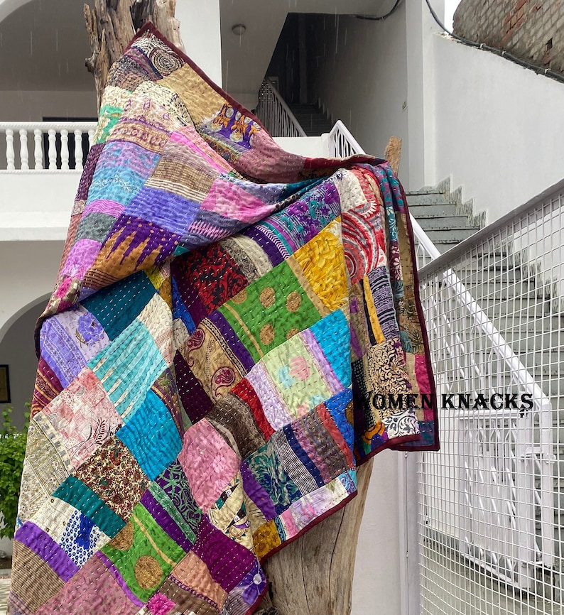 Pu&ograve; includere: Una trapunta patchwork composta da vari quadrati di tessuto colorato con motivi diversi, inclusi disegni floreali, paisley e geometrici. La trapunta presenta cuciture kantha visibili e un bordo rosso scuro. Il testo WOMEN KNACKS &egrave; stampato.