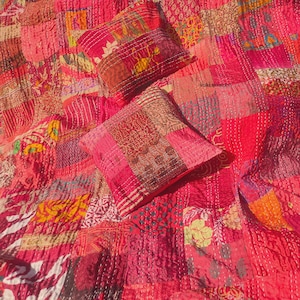 Könnte beinhalten: Eine Patchwork-Steppdecke und zwei passende Wurfkissen in Rot- und Rosatönen. Die Steppdecke und die Kissen bestehen aus verschiedenen Stoffresten, die mit sichtbarem weißen Faden zusammengenäht wurden. Das Gesamtdesign ist eine Mischung aus Mustern und Texturen.