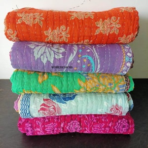 May include: A stack of five kantha quilts in various colors and patterns. The quilts are made of cotton and are hand-stitched. The top quilt is orange with a floral pattern, the second is purple with a paisley pattern, the third is green with a floral pattern, the fourth is light blue with a floral pattern, and the bottom quilt is pink with a floral pattern. The quilts are all folded in half and stacked on top of each other. The word "WOMENKNACKS" is visible on the second quilt.