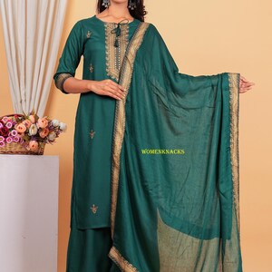 May include: A teal green kurta set with gold embroidery and a matching dupatta. The set features a long-sleeved kurta, palazzo pants, and a sheer dupatta. The kurta has a round neckline and a tie-up detail at the front. The palazzo pants have a wide leg and a gold embroidered border. The dupatta is sheer and has a gold embroidered border.