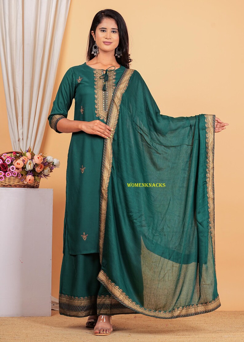 May include: A teal green kurta set with gold embroidery and a matching dupatta. The set features a long-sleeved kurta, palazzo pants, and a sheer dupatta. The kurta has a round neckline and a tie-up detail at the front. The palazzo pants have a wide leg and a gold embroidered border. The dupatta is sheer and has a gold embroidered border.