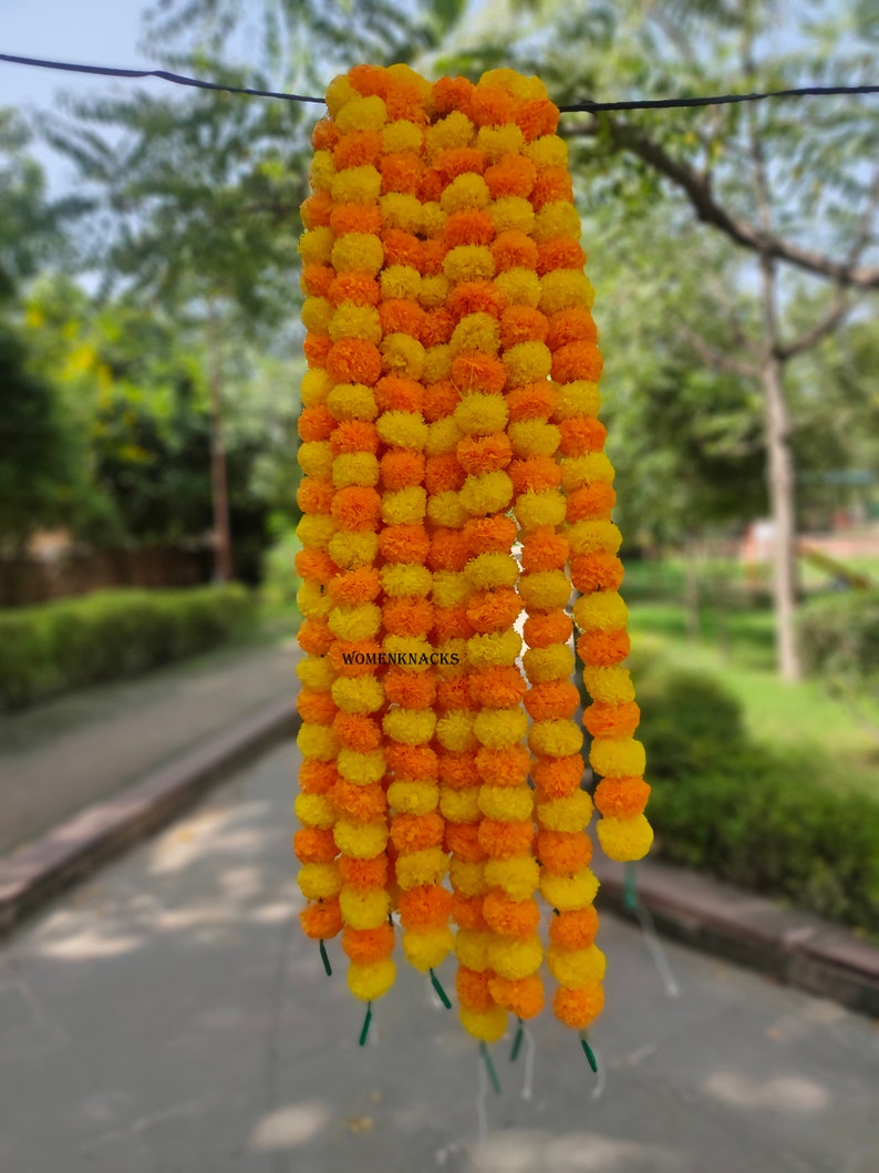 K&ouml;nnte beinhalten: Eine Girlande aus gelben und orangefarbenen Margeriten-Bl&uuml;tenkugeln, die aneinandergereiht sind und an einem Draht h&auml;ngen. Die Girlande ist etwa 90 cm lang und besteht aus k&uuml;nstlichen Blumen.