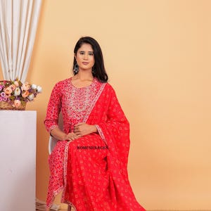 May include: A red and white patterned dress with a matching dupatta. The dress has a round neckline and three-quarter sleeves. The dupatta has a white border. The model is wearing the dress and dupatta and is sitting on a chair. The text "WOMENKNACKS" is visible on the dupatta.