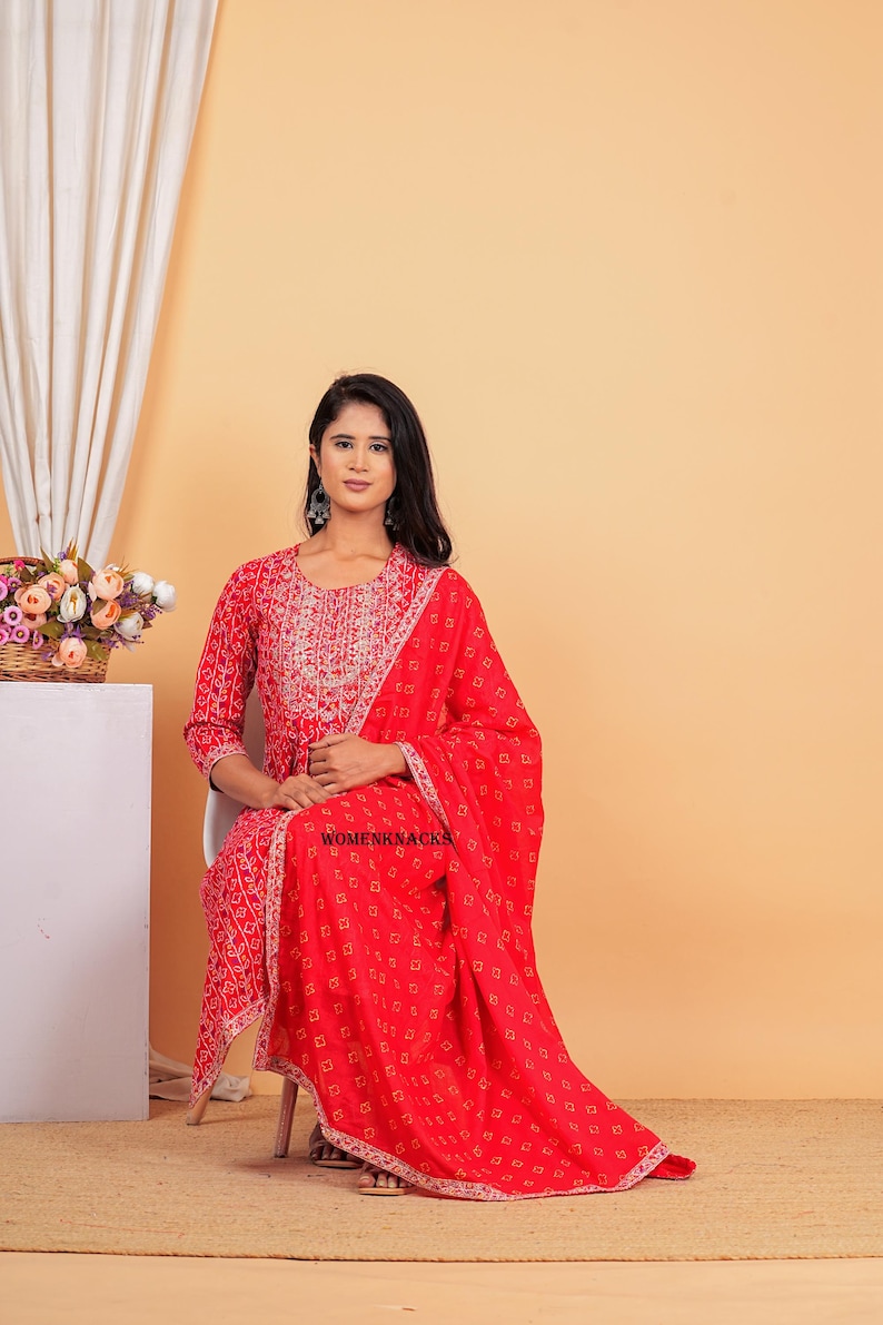 May include: A red and white patterned dress with a matching dupatta. The dress has a round neckline and three-quarter sleeves. The dupatta has a white border. The model is wearing the dress and dupatta and is sitting on a chair. The text "WOMENKNACKS" is visible on the dupatta.