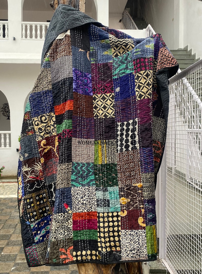 Pu&ograve; includere: Una trapunta patchwork caratterizzata da una variet&agrave; di pannelli rettangolari in tessuto con colori e motivi diversi, tra cui nero, blu, verde e oro. Il tessuto &egrave; cucito con un filo bianco visibile e riporta il testo WOMENK MAGIC.