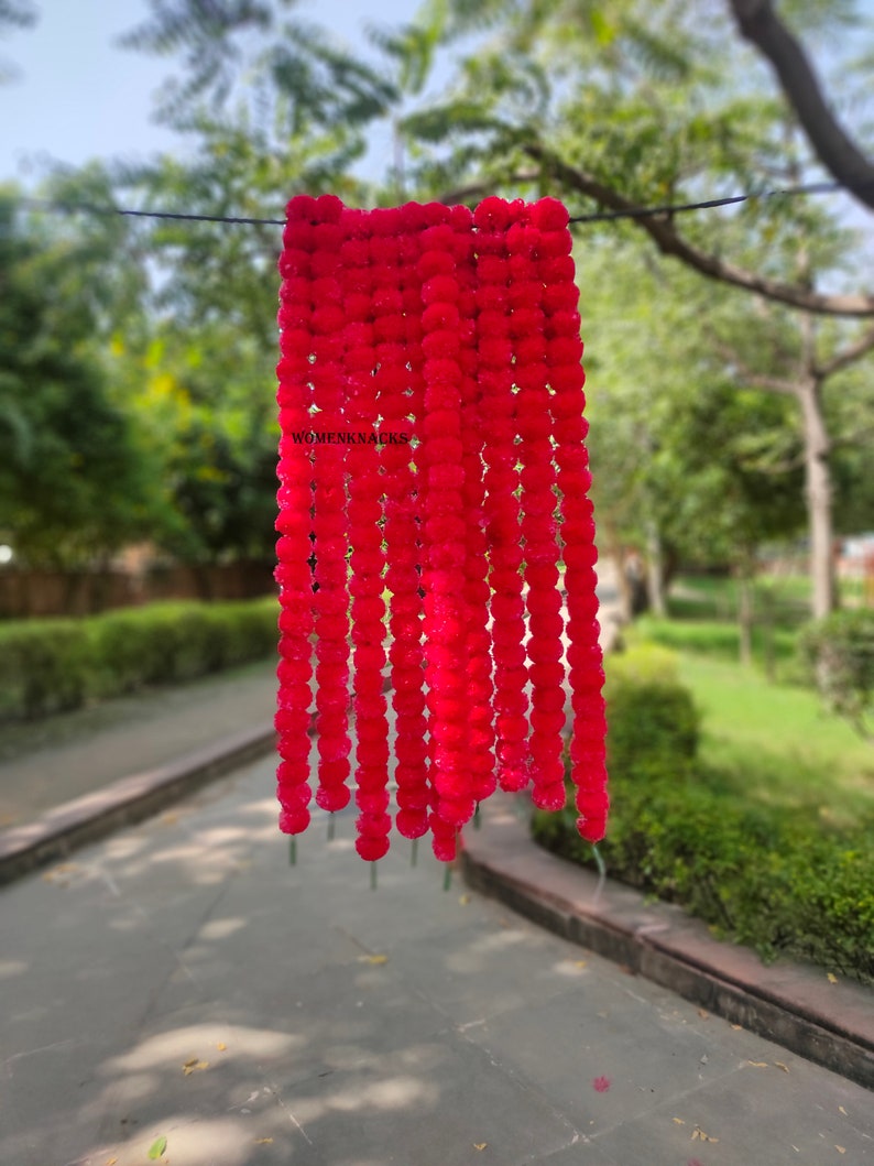 K&ouml;nnte beinhalten: Eine Girlande aus roten k&uuml;nstlichen Bl&uuml;tenpompons, die an einem Draht h&auml;ngen. Die Pompons bestehen aus weichem, flauschigem Material und sind in einer kaskadierenden Anordnung angeordnet. Der Text "WOMENKNACKS" ist auf der Girlande sichtbar.
