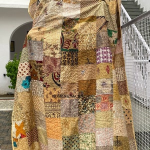 May include: A patchwork quilt made of various fabrics in shades of brown, beige, and cream. The quilt is made up of squares of different patterns and colors, and is quilted with a simple stitch. The quilt is hanging on a tree branch.