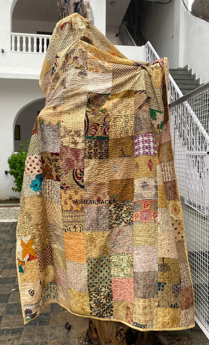 May include: A patchwork quilt made of various fabrics in shades of brown, beige, and cream. The quilt is made up of squares of different patterns and colors, and is quilted with a simple stitch. The quilt is hanging on a tree branch.