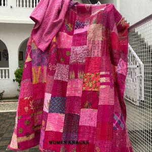 May include: A pink kantha quilt with a patchwork design. The quilt is made of various fabrics in shades of pink, purple, and blue. The quilt is hand-stitched and has a unique, rustic look. The quilt is folded and draped over a wooden post. The text "WOMEN KNACKS" is visible in the bottom right corner of the image.