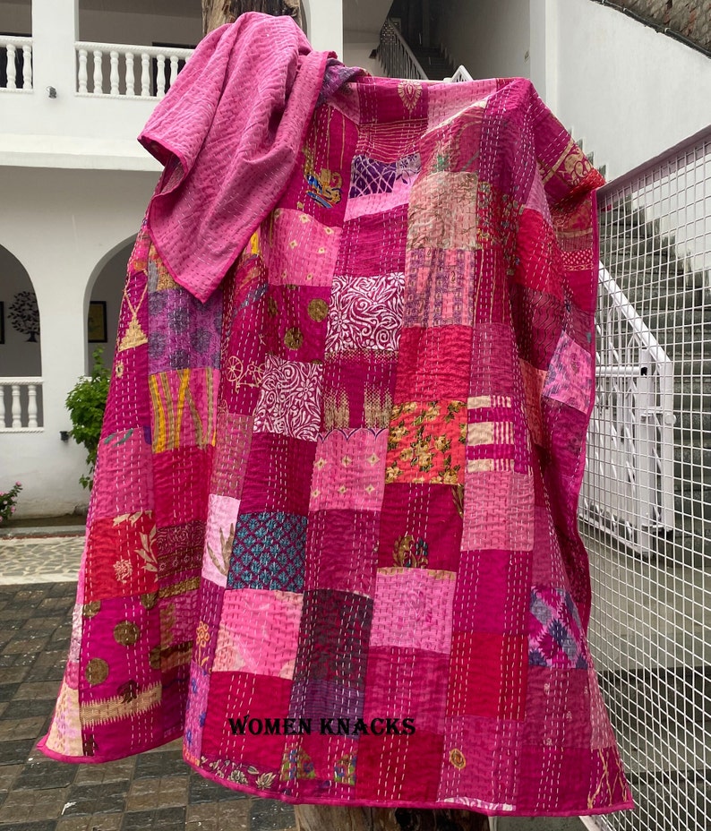 May include: A pink kantha quilt with a patchwork design. The quilt is made of various fabrics in shades of pink, purple, and blue. The quilt is hand-stitched and has a unique, rustic look. The quilt is folded and draped over a wooden post. The text "WOMEN KNACKS" is visible in the bottom right corner of the image.