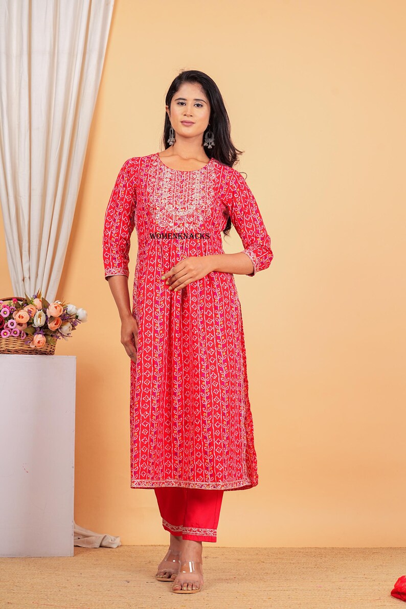 May include: A red and white printed kurta with a gold embroidered neckline and a matching pair of red pants. The kurta has a gathered waist and a long, flowing silhouette. The pants are slim-fitting and have a gold embroidered trim at the hem.