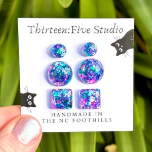 May include: A set of six stud earrings on a white display card labelled Thirteen:Five Studio and Handmade in the NC Foothills. The earrings feature a purple, blue, and teal glitter resin finish in small round, large round, and square shapes.