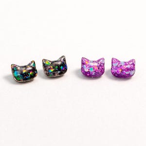 May include: Four cat-shaped stud earrings. Two are black with iridescent glitter, and two are purple with glitter. The earrings are small and have a smooth, rounded shape. They are set against a white background.