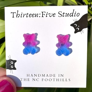 May include: A pair of stud earrings shaped like gummy bears with a gradient of pink and blue, mounted on a white display card featuring the text Thirteen:Five Studio and HANDMADE IN THE NC FOOTHILLS, along with black cat illustrations.