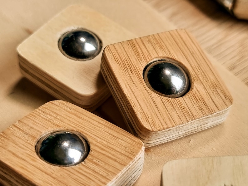 May include: Close-up of three wooden squares, each with a recessed, shiny metal ball. The squares have a natural wood grain finish and are arranged on a light-colored wooden surface. The design is simple and modern.