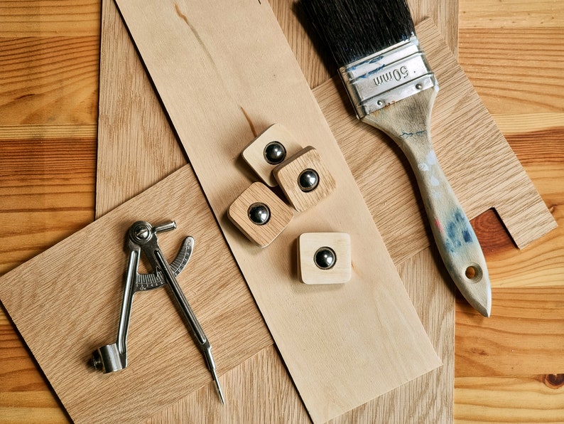 May include: An overhead shot of woodworking tools and materials. Includes a paintbrush, a compass, wooden squares with metal spheres, and various wood pieces. The paintbrush has a 50mm marking on the metal. The wood has a natural grain.
