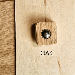 May include: A square oak cabinet knob with a circular, silver-colored metal insert. The knob is set against a light-colored wooden surface, with the word "OAK" printed in black below it.