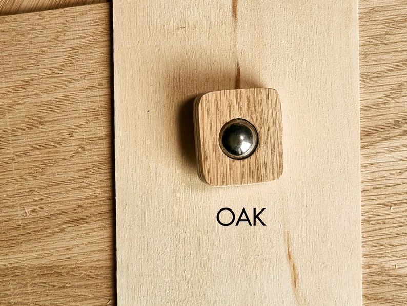 May include: A square oak cabinet knob with a circular, silver-colored metal insert. The knob is set against a light-colored wooden surface, with the word "OAK" printed in black below it.
