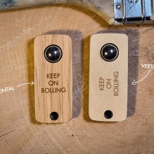 Op de afbeelding: Twee houten deegrollerhandvatten, één licht en één donker, met een metalen kogellager en de woorden "KEEP ON ROLLING" gegraveerd. De handvatten zijn rechthoekig met een gat aan de onderkant. Eén is gemarkeerd met "HORIZONTAL" en de andere met "VERTICAL".