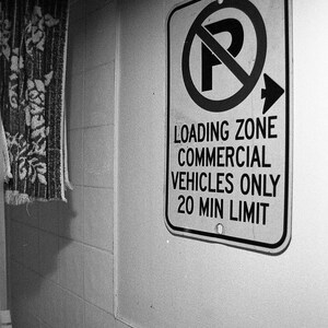 Może przedstawiać: Czarno-białe zdjęcie znaku parkingowego z napisem "Loading Zone Commercial Vehicles Only 20 Min Limit". Znak jest prostokątny z czarną obwódką i symbolem zakazu parkowania. Jest zamontowany na białej kafelkowej ścianie.