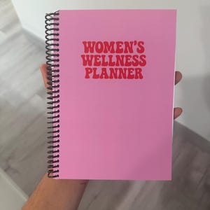 以下が含まれることがあります： 赤い文字で「WOMEN'S WELLNESS PLANNER」と書かれたピンク色のリング綴じプランナー。白い背景を背景に、人の手でプランナーが持たれています。表紙は無地のピンク色です。