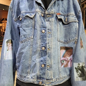 May include: A blue denim jacket with multiple patches, including a portrait of a person with blue eyes and a red and white design. The jacket has a button closure and five buttons down the front.