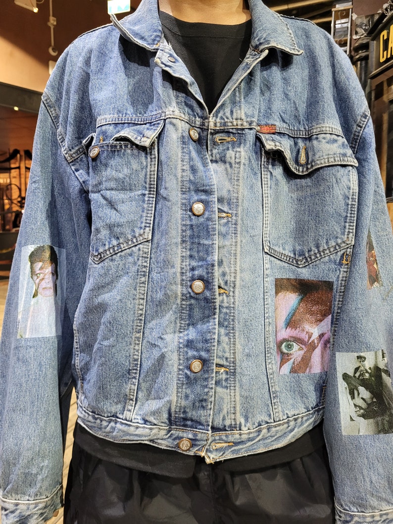 May include: A blue denim jacket with multiple patches, including a portrait of a person with blue eyes and a red and white design. The jacket has a button closure and five buttons down the front.