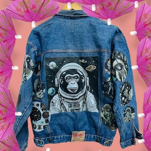 Może przedstawiać: Niebieska dżinsowa kurtka z dużą naszywką z tyłu przedstawiającą szympansa w kosmicznym skafandrze. Kurtka ma kilka mniejszych naszywek z kosmicznymi wzorami.